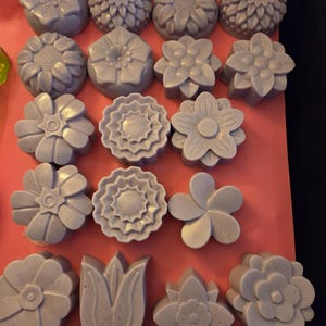 Puede incluir: Una colección de dieciocho pastillas de jabón de color morado claro con forma de flor, dispuestas en filas sobre una superficie rosa. Cada jabón presenta un diseño floral único, incluyendo girasoles, margaritas y tulipanes, con una textura moteada.