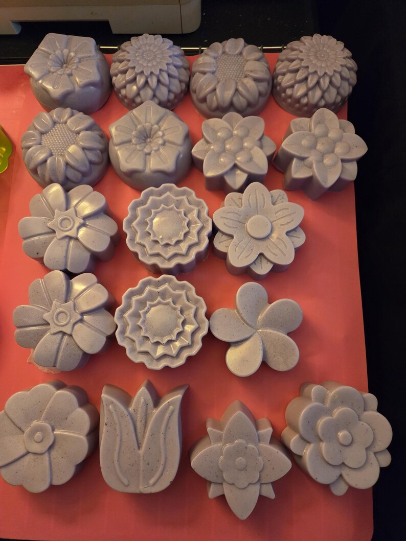 May include: A collection of eighteen light purple, flower-shaped soap bars arranged in rows on a pink surface. Each soap features a unique floral design, including sunflowers, daisies, and tulips, with a subtle speckled texture throughout.