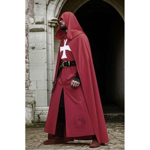 Op de afbeelding: Een bordeauxrode cape met capuchon en een wit kruisembleem op de borst. De cape heeft een split aan de voorkant, een zwarte riem en wordt gedragen met donkere laarzen. De afbeelding is geplaatst tegen een stenen gebouw met een boogvormige deuropening.