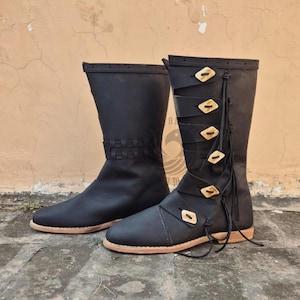 May include: A pair of black leather boots. One boot features decorative wooden buttons and straps, while the other has a woven design at the top. Both boots have a light brown sole.
