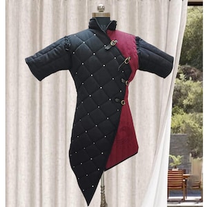 May include: Black and burgundy quilted medieval gambeson with short sleeves. The gambeson features a high collar, diamond-patterned stitching, and decorative metal clasps. The garment is displayed on a mannequin against a neutral background.