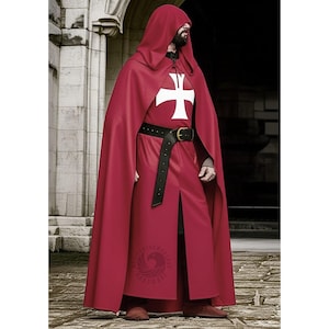Op de afbeelding: Een rode cape met capuchon en bijpassende tuniek met een wit kruisembleem. De outfit bevat een zwarte riem met een gouden gesp. De cape is lang en vloeiend, en de tuniek heeft een split aan de voorkant. Middeleeuws kostuum.