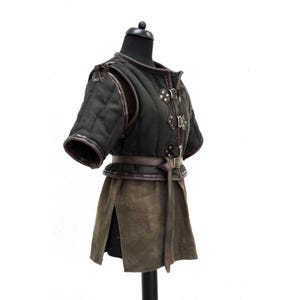 May include: A dark green medieval-style doublet with short sleeves, brown leather trim, and decorative metal clasps. The garment features a matching belt and a split skirt, displayed on a black mannequin.