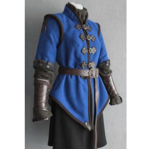 Puede incluir: Una chaqueta azul de estilo medieval con detalles en cuero marrón y un cinturón a juego. La chaqueta presenta cierres decorativos, hombreras y protectores de brazo de cuero. La parte inferior de la chaqueta tiene un diseño angular único y se combina con una falda negra.