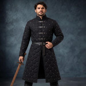 May include: A black quilted long coat featuring a diamond pattern with small silver studs at each intersection. The coat has a high collar, long sleeves, a front closure with four silver metal buckles, and a black leather belt at the waist.