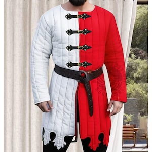 Könnte beinhalten: Gesteppte Tunika in Weiß und Rot, vertikal geteilt. Sie hat schwarze Lederriemen mit goldenen Schnallen, einen schwarzen Gürtel und dekorative Kanten. Das langärmlige Kleidungsstück ist für historische Nachstellungen oder Kostüme geeignet.