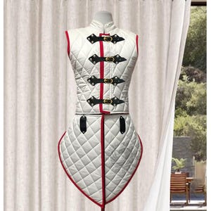 May include: White quilted vest and skirt set with red trim and black leather straps with gold buckles. The diamond-patterned fabric is displayed on a mannequin. The outfit is designed for historical reenactment or costume use.