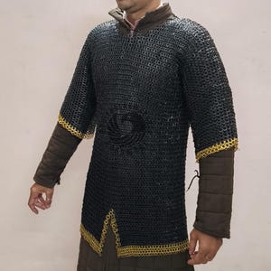 Puede incluir: Camisa de cota de malla negra con ribete dorado en las mangas y el dobladillo. La camisa tiene mangas largas y se usa sobre una prenda acolchada marrón. La cota de malla está hecha de pequeños anillos de metal entrelazados.