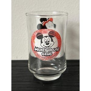 May include: Clear glass tumbler featuring a Mickey Mouse Club logo with Mickey Mouse and Minnie Mouse illustrations. The logo is a red circle with white text and a black and white Mickey Mouse image. The glass is on a black surface.