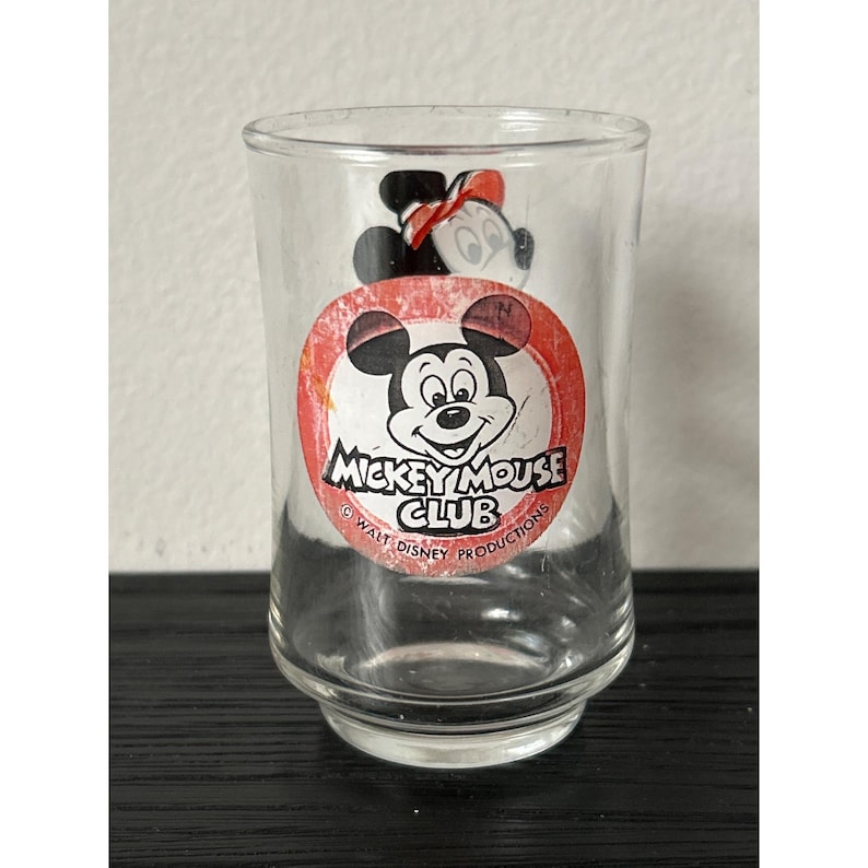 May include: Clear glass tumbler featuring a Mickey Mouse Club logo with Mickey Mouse and Minnie Mouse illustrations. The logo is a red circle with white text and a black and white Mickey Mouse image. The glass is on a black surface.