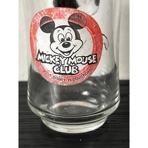 May include: A clear glass with a Mickey Mouse Club logo. The logo features Mickey Mouse's face in a red circle with black ears and the text "Mickey Mouse Club" in white. The glass is a vintage collectible.