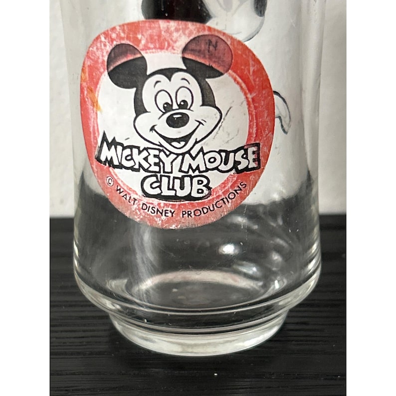 May include: A clear glass with a Mickey Mouse Club logo. The logo features Mickey Mouse's face in a red circle with black ears and the text "Mickey Mouse Club" in white. The glass is a vintage collectible.