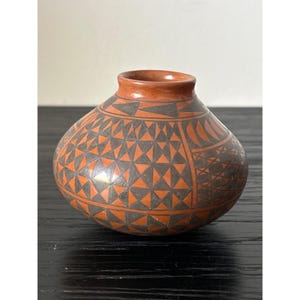 May include: A small, round, terracotta-coloured ceramic vase with a narrow neck. The vase features a geometric pattern of black triangles and shapes over an orange background. The design covers the body of the vase, creating a striking visual contrast.