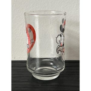 May include: Clear glass tumbler featuring a heart-shaped design with the text "Mickey Mouse" and a cartoon image of Mickey Mouse. The glass has a rounded base and a straight, cylindrical body. The design includes red, black, and white colors.