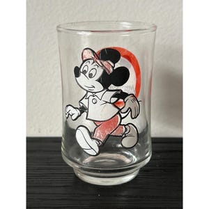 May include: Clear glass tumbler featuring a running cartoon character in a white shirt, red pants, and a pink bow. The character is black and white with a red circle in the background. A vintage Disney collectible.