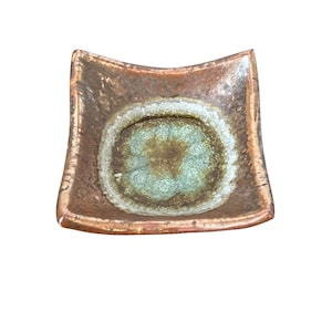 May include: Square ceramic dish with a copper-colored exterior and a central design in shades of green, brown, and white. The dish has a slightly curved shape and a glossy finish, suitable for serving small items.