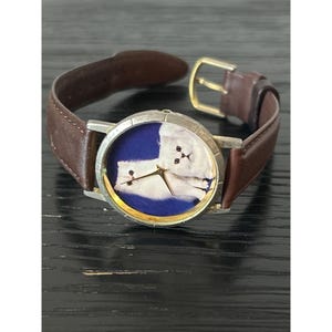 May include: A wristwatch with a brown leather band and a silver-tone case. The watch face features a blue background with a white cat illustration. The watch has gold-tone hands and a gold-tone buckle.