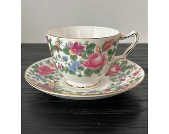 Può includere: Servizio da tè bianco con un motivo floreale con rose rosa, fiori blu e gialli e foglie verdi. La tazza e il piattino hanno finiture dorate.