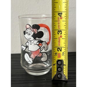 May include: Clear glass tumbler featuring a vintage Mickey Mouse design. The cartoon character is depicted in a walking pose, wearing a hat, shirt, and pants in red, white, and black. The tumbler is approximately 3 inches tall.