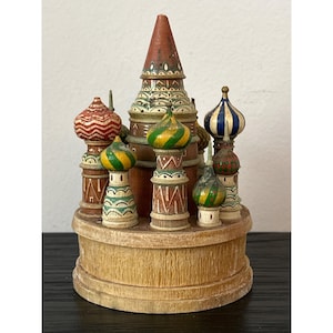 May include: A wooden miniature of a Russian Orthodox church with multiple colorful onion domes. The central tower is reddish-brown, and the domes are painted in various colors, including red, green, blue, and yellow. The base is a light brown, circular structure.