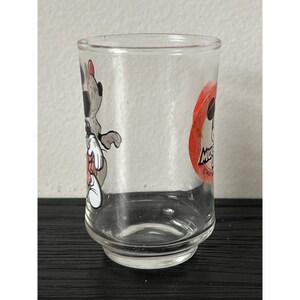 May include: A clear glass tumbler featuring cartoon character Mickey Mouse. The tumbler has a cylindrical shape with a slightly flared base. Mickey Mouse is depicted in a red and white circle on the side of the glass.
