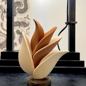 May include: A wooden sculpture of a stylized flower with layered petals in shades of brown and cream. The sculpture is mounted on a small, round base and sits on a dark surface. The background includes a black frame and a patterned wall.