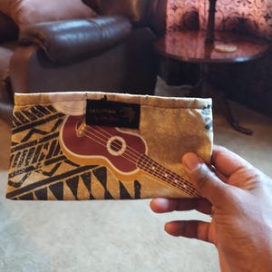 May include: A rectangular fabric case with a ukulele design. The case features a brown ukulele graphic on a tan background with black tribal patterns. The text "La Lumiere" is visible on a black label.