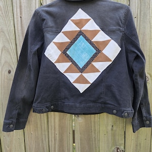 May include: A dark gray denim jacket with a quilt-style design on the back. The quilt pattern features a central turquoise square surrounded by brown, white, and black geometric shapes. The jacket has button closures and pockets.