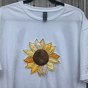May include: A white t-shirt with an appliqued sunflower design. The sunflower has a brown centre, yellow and gold petals, and a patterned accent. The shirt is hanging on a black hanger against a wooden background.