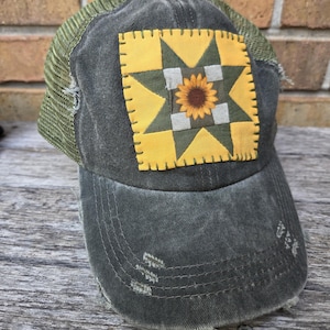 May include: Distressed grey baseball cap with a green mesh back. The front features a patch with a yellow and green star design, and a sunflower in the centre. The cap has a worn, vintage look.