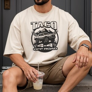 May include: Cream-colored t-shirt featuring a black graphic of a truck, mountains, and the words "TACO 4x4 OFF ROAD." The shirt has short sleeves and a relaxed fit. The person is wearing khaki shorts and a watch.