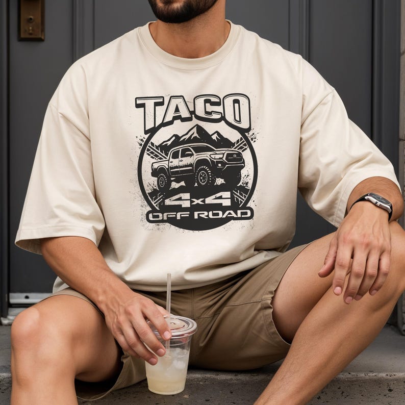 May include: Cream-colored t-shirt featuring a black graphic of a truck, mountains, and the words "TACO 4x4 OFF ROAD." The shirt has short sleeves and a relaxed fit. The person is wearing khaki shorts and a watch.