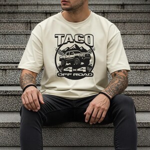 May include: Cream-colored t-shirt featuring a black graphic design. The design includes the word "TACO" above a mountain range and a 4x4 off-road vehicle. Below the vehicle are the words "4x4 OFF ROAD".