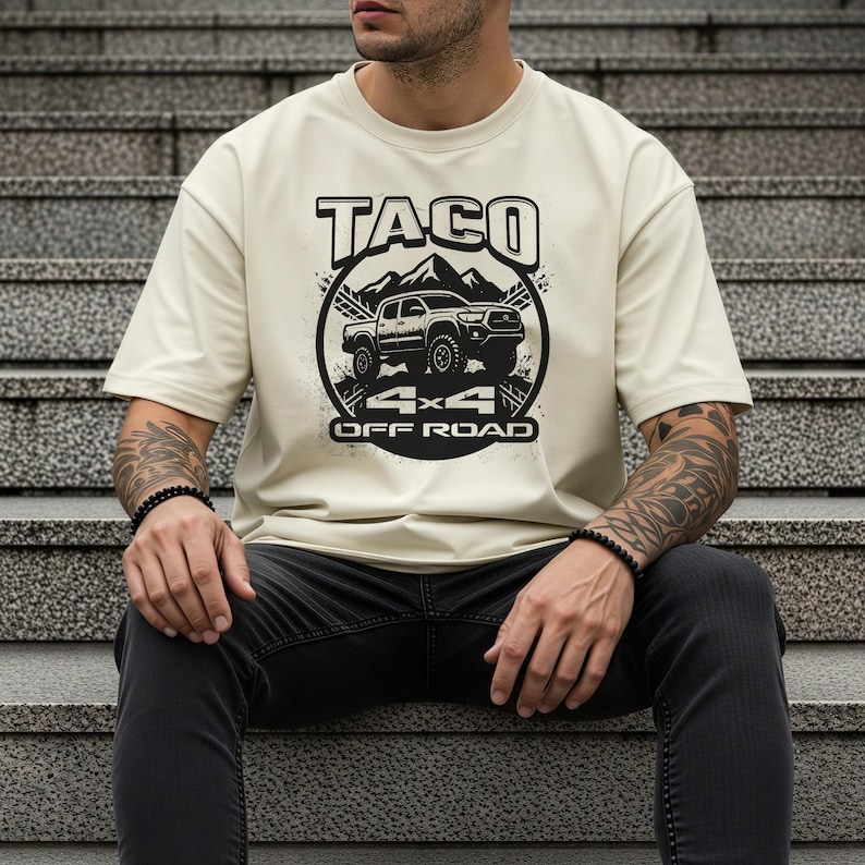 May include: Cream-colored t-shirt featuring a black graphic design. The design includes the word "TACO" above a mountain range and a 4x4 off-road vehicle. Below the vehicle are the words "4x4 OFF ROAD".