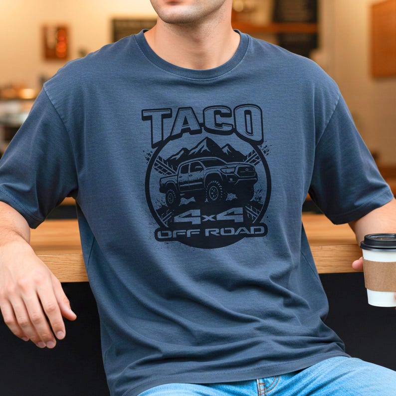 May include: A blue-grey t-shirt featuring a black graphic design. The design includes the word "TACO" above an illustration of a truck in front of mountains, with "4x4 OFF ROAD" below. The shirt has a crew neck and short sleeves.