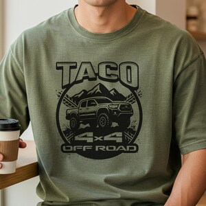 May include: Olive green t-shirt with a black graphic featuring the word "TACO" above an off-road truck illustration, mountains, and the text "4x4 OFF ROAD". The shirt has a crew neck and short sleeves.