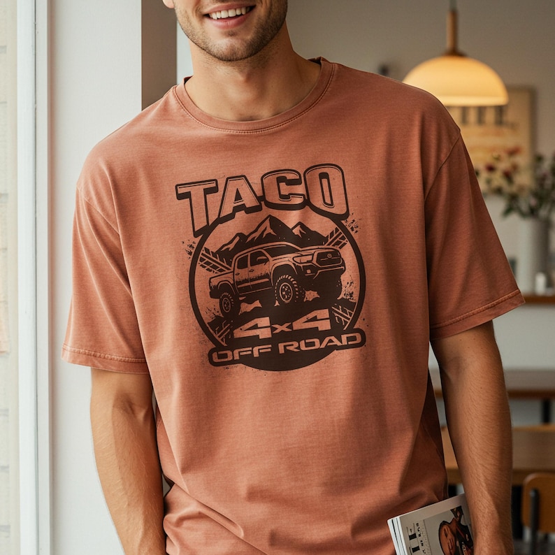 May include: A rust-colored t-shirt featuring a graphic of a truck with the text "TACO 4x4 OFF ROAD" in a circular design. The design includes a mountain range and a stylized image of the truck. The shirt has short sleeves and a crew neck.