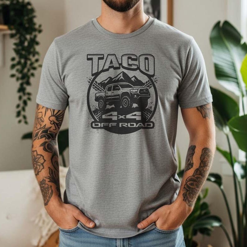 May include: A gray t-shirt with a black graphic design featuring the word "TACO" above an illustration of a truck, mountains, and the text "4x4 OFF ROAD". The shirt is short-sleeved.