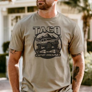 May include: Olive green t-shirt with a black graphic design. The design features the word "TACO" above an illustration of a truck, mountains, and the text "4x4 OFF ROAD". The shirt is made of a soft material.
