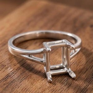 Op de afbeelding: Een zilveren ringzetting met een rechthoekige kop met vier pootjes, ontworpen om een edelsteen vast te houden. De band heeft een gespleten ontwerp dat aansluit op de zetting, wat zorgt voor een gepolijste metalen afwerking.