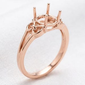 Peut inclure: Monture de bague de fiançailles en or rose, avec quatre griffes pour une pierre ronde. La bague présente une monture fendue et des détails décoratifs sur les épaules. Fond blanc.