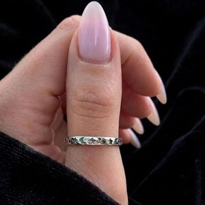 May include: A thin silver band ring featuring a repeating pattern of small engraved stars and crescent moons around the entire circumference of the metal surface. The ring has a polished finish and a flat, uniform width throughout its design.