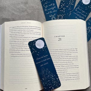 May include: Dark blue bookmarks with white stars and a full moon design. Each bookmark features a quote in white text. One bookmark is in a book, with others nearby.