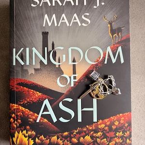 Puede incluir: Un libro de Sarah J. Maas, "Kingdom of Ash", con una portada que muestra un castillo, un ciervo y un llavero. Pertenece a la serie "Throne of Glass", publicado por Bloomsbury.