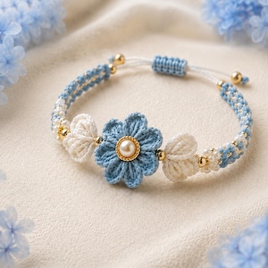 May include: A woven macrame bracelet featuring a central blue flower with a pearl center, flanked by two smaller white flower motifs. The band is crafted from blue and white cord, accented with small gold-toned beads and an adjustable sliding knot closure.
