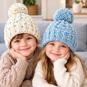 May include: Two knit beanies with pom-poms. One beanie is cream-colored with blue and yellow speckles, and the other is light blue with white and yellow speckles. Both hats feature a thick ribbed texture and a large, round pom-pom on top.