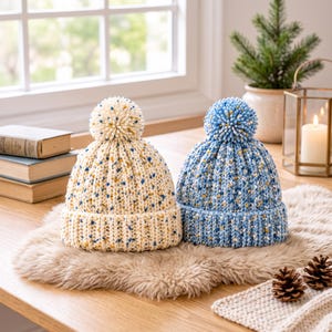 May include: Two knit beanies with pom-poms, one in cream with blue and yellow flecks and one in blue with white and yellow flecks. Both hats feature a thick ribbed cuff and a textured, chunky knit pattern, showcasing a speckled yarn design.