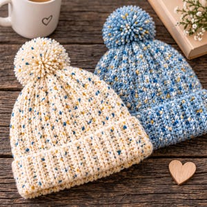 May include: Two knit beanies with pom-poms, one in cream and one in blue, both featuring a speckled pattern of small blue and yellow flecks throughout the yarn. Each hat has a thick ribbed cuff and a textured knit construction with a round pom-pom.