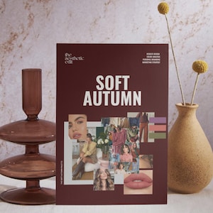 May include: A burgundy magazine titled "Soft Autumn" with a collage of images and text. A decorative glass candle holder and a textured vase with dried flowers are on either side of the magazine. The magazine is a style guide.