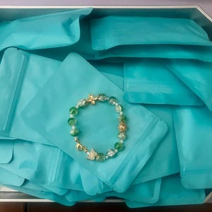 May include: A beaded bracelet with green and gold accents rests on a pile of turquoise pouches inside a silver tray. The bracelet features clear and green beads, gold-colored spacers, and a charm. The pouches are sealed and ready for packaging.